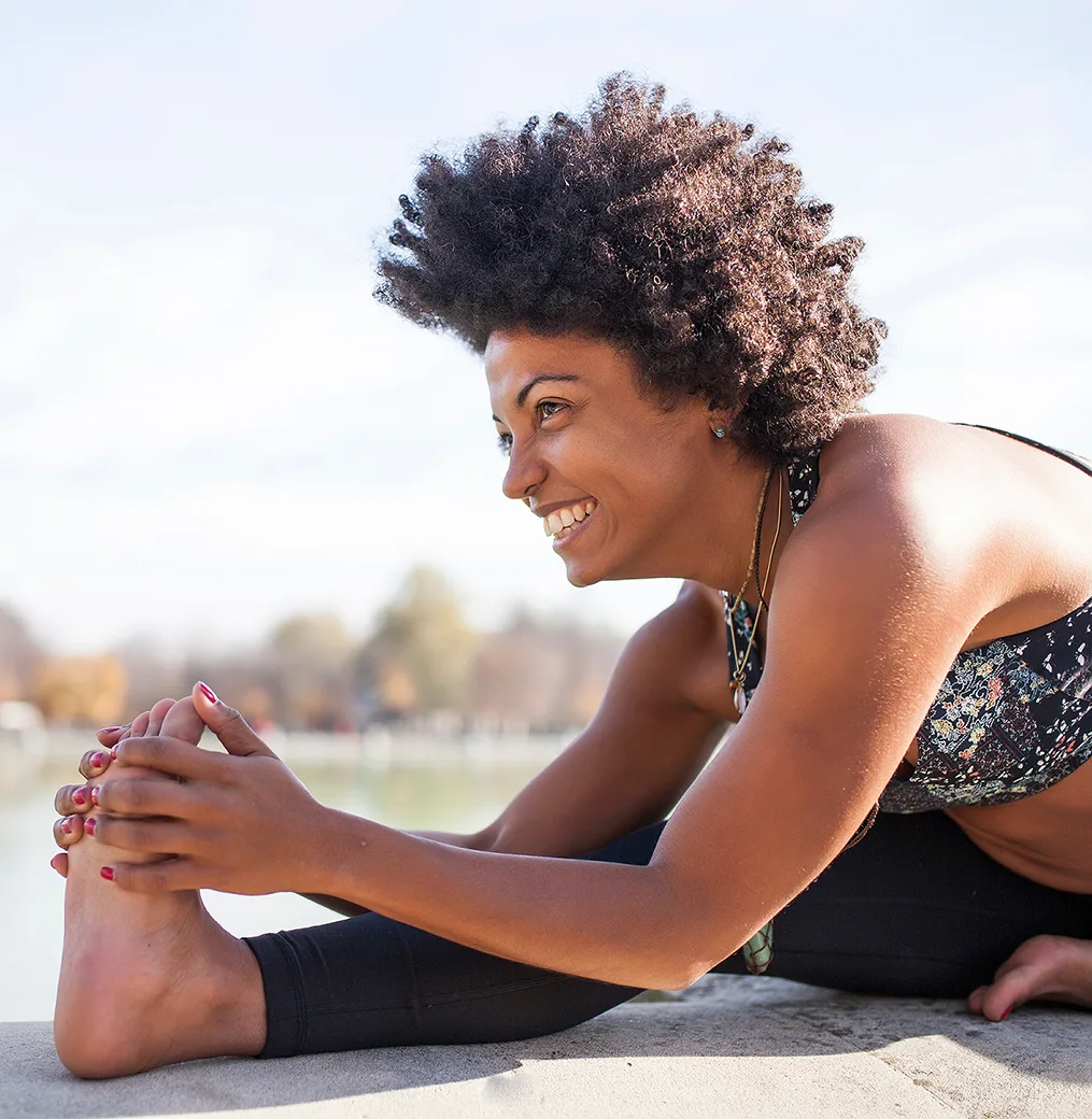 A smiling woman stretching forward to her toes beside a lake